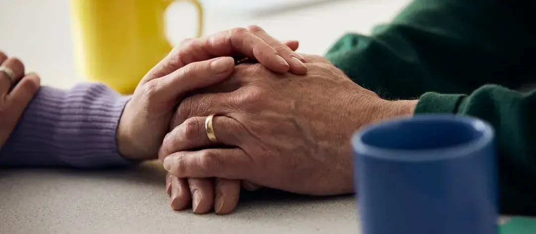 One persons hand laid over another person's hand signifying support and connection. The presence of two mugs suggests a comforting setting.