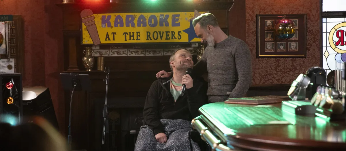 Coronation Street character, Paul Foreman, looks up from his chair to another character as they smile at one another adoringly. He holds a microphone in a pub setting, and a sign in the background reads 'Karaoke at the Rovers.'