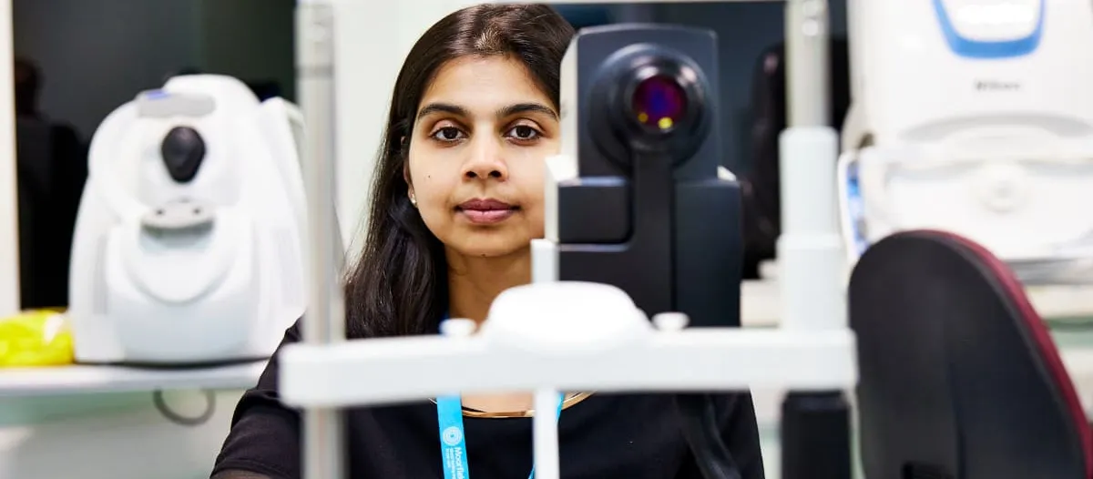 A young woman sits at an eye examination station in a clinical setting wearing a Moorfields Eye Charity lanyard.